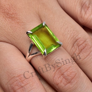 Puede incluir: Un anillo de plata con una gran piedra preciosa verde peridoto de talla esmeralda. El anillo está engastado en un diseño simple y elegante.