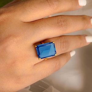 Könnte beinhalten: Ein goldfarbener Ring mit einem großen, rechteckigen, leuchtend blauen Edelstein. Die Fassung des Rings hat kleine goldfarbene Akzente an jeder Ecke des Edelsteins. Der Ring wird an einer Hand mit weißem Nagellack getragen.