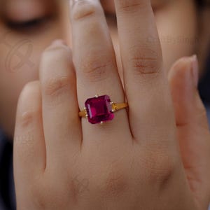 Peut inclure: Une bague en or ornée d'une grosse pierre précieuse rouge foncé de forme rectangulaire. La bague est sertie sur un anneau en or et est présentée sur un doigt. L'arrière-plan est flou.