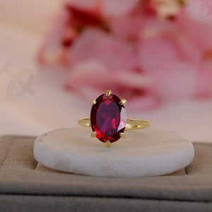 May include: A gold ring with a large, oval-cut, deep red gemstone. The ring is set on a white stone, atop a gray box, with pink flowers in the background. The ring is a piece of jewelry.