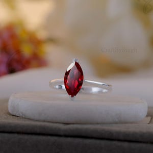 Op de afbeelding: Een zilveren ring met een marquise-geslepen, dieprode edelsteen. De ring staat op een witte, cilindrische basis, met een vage achtergrond van bloemen en stof. De ring heeft een slanke, gepolijste band.