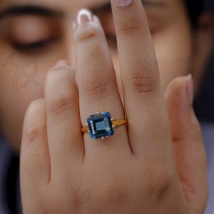 May include: A gold ring with a large, rectangular, deep blue gemstone. The ring has decorative side details. The ring is shown on a person's finger.