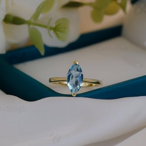 May include: A marquise-cut, light blue gemstone ring with a gold band. The ring is displayed in a teal box on a white satin cloth, with white flowers in the background. The ring is the focal point of the image.