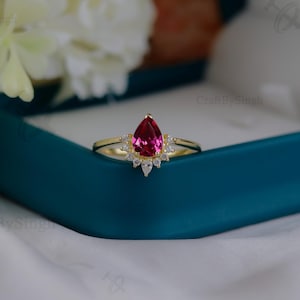 May include: A gold ring with a pear-shaped, vibrant pink gemstone as the centerpiece. The ring is accented with smaller, clear gemstones around the main stone. The ring is displayed in a teal jewelry box, with white flowers in the background.