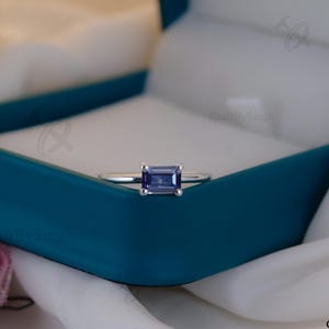 May include: A silver ring with a rectangular, light blue gemstone. The ring is set in a teal jewelry box with a white interior. A pink rose and white fabric are in the background.