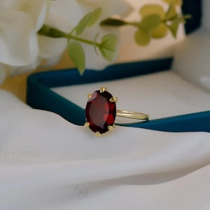 Puede incluir: Un anillo de oro con una gran piedra preciosa ovalada de color rojo oscuro. El anillo se encuentra sobre una tela blanca y una caja verde azulado, con flores blancas en el fondo. El anillo es una joya.