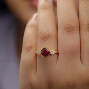 Könnte beinhalten: Ein goldener Ring mit einem großen, quadratisch geschliffenen, tiefroten Edelstein als Mittelpunkt, flankiert von zwei kleineren, klaren, rautenförmigen Steinen. Der Ring wird an einer Hand getragen, um das Design und die Farbe hervorzuheben.