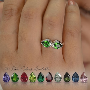Könnte beinhalten: Ein silberner Ring mit zwei birnenförmigen, leuchtend grünen Edelsteinen. Der Ring wird auf einer Hand gezeigt, mit einer Auswahl anderer Edelsteinfarben darunter. Der Text auf dem Bild lautet "20+ Stone Colors Available."