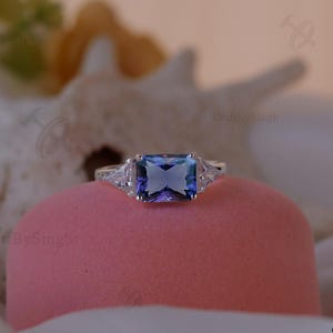May include: A silver ring featuring a large, rectangular, blue-purple gemstone, with two smaller, triangular, clear gemstones either side. The ring is displayed on a pink surface, with a blurred white and yellow background.