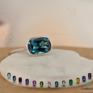 May include: A silver ring with a large, rectangular, teal gemstone. The ring sits on a white, textured surface, with a selection of smaller, colourful gemstones displayed below. The ring is a piece of jewellery.