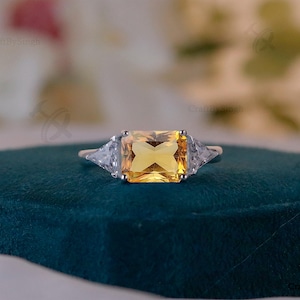 May include: A silver ring with a large, rectangular, yellow gemstone as the centerpiece. Two smaller, triangular, clear gemstones flank the main stone. The ring is set on a dark teal surface, with a blurred background.