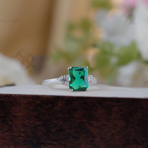 Könnte beinhalten: Ein silberner Ring mit einem großen, smaragdgeschliffenen grünen Edelstein als Mittelpunkt. Der Ring hat kleinere, klare Steine auf beiden Seiten des Hauptsteins. Der Ring wird auf einer dunkelbraunen Oberfläche präsentiert.