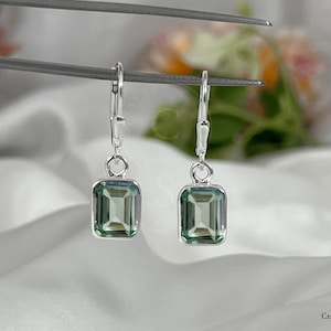 May include: A pair of silver drop earrings featuring rectangular, emerald-cut, green gemstones. The earrings hang from lever-back closures. The background is a soft, white fabric with a blurred floral arrangement.