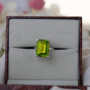 May include: A silver ring with a large, rectangular, light green gemstone, set in a brown jewelry box. The ring is centered in the box, with the gemstone prominently displayed. The box is open, revealing the ring against a neutral background.