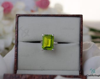 May include: A silver ring with a large, rectangular, light green gemstone, set in a brown jewelry box. The ring is centered in the box, with the gemstone prominently displayed. The box is open, revealing the ring against a neutral background.