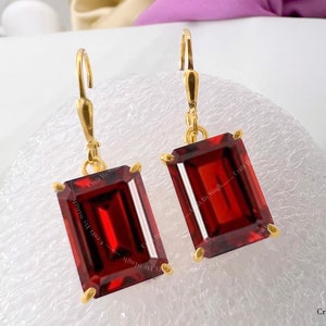 Könnte beinhalten: Ein Paar goldene Ohrhänger mit roten rechteckigen Edelsteinen. Die Ohrringe haben ein schlichtes Design und sind perfekt für den Alltag.