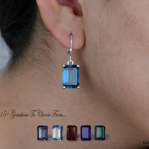 Puede incluir: Un pendiente de plata con una gema azul rectangular se lleva en una oreja. El pendiente es de estilo palanca. La imagen también muestra una selección de gemas en varios colores y el texto "15+ Gemstone To Choose From.."