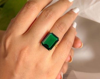 Op de afbeelding: Een gouden ring met een grote, smaragdgeslepen, diepgroene edelsteen. De ring heeft een gouden zetting en de edelsteen is rechthoekig. De ring zit aan een hand met witte nagellak.