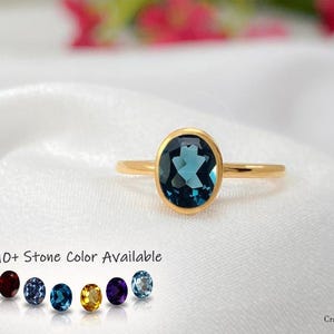 Op de afbeelding: Een gouden ring met een centrale ovaal geslepen donkerblauwe edelsteen in een gladde zetting. Onder de ring staat de tekst 10+ Stone Color Available naast zes kleine ronde edelsteenvoorbeelden in rode, blauwe, gele en paarse tinten.