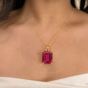 Puede incluir: Un collar de cadena dorada con un gran colgante rectangular de piedra preciosa magenta en talla esmeralda, montado en un engaste de garras dorado. El colgante cuelga de una fina cadena dorada. El texto CraftBySingh es visible en la esquina inferior derecha.