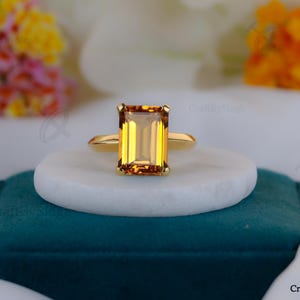 May include: A gold ring with a large, rectangular, yellow-orange gemstone. The ring is set on a white, round surface, atop a teal velvet box. The background features blurred flowers.