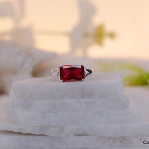 以下が含まれることがあります： 大きな長方形の濃い赤色の宝石を特徴とするシルバーリング。2つの小さな三角形のクリアストーンがメインストーンを挟んでいます。リングは白い層状の表面にセットされています。