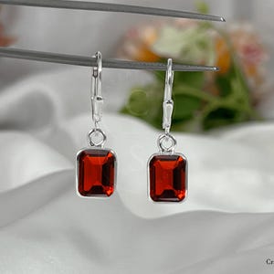 Peut inclure: Boucles d'oreilles argentées avec des pierres précieuses rectangulaires rouge foncé. Les boucles d'oreilles ont une fermeture à levier et sont présentées sur un fond blanc et soyeux. Les pierres sont d'une couleur vive et saturée.