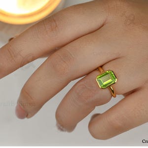 May include: A gold ring with a rectangular, light green gemstone. The ring has a thin, polished gold band, and the gemstone is set in a bezel setting. The ring is displayed on a finger, against a soft, neutral background.