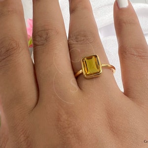 May include: A gold ring with a large, rectangular, yellow gemstone. The ring is on a person's finger.