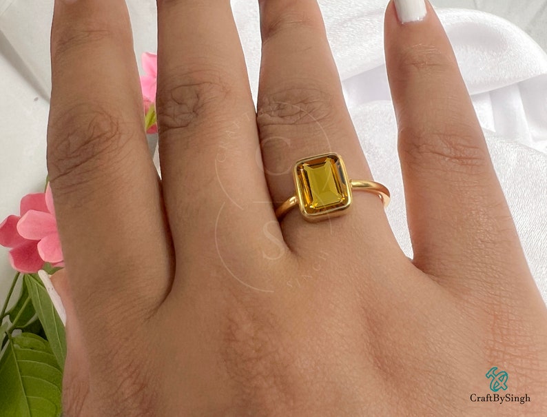 May include: A gold ring with a large, rectangular, yellow gemstone. The ring is on a person's finger.