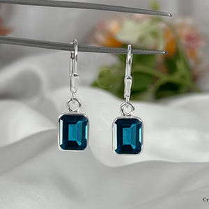 May include: A pair of silver leverback earrings featuring rectangular, emerald-cut dark blue stones. The earrings are suspended from thin metal bars, showcasing a polished silver setting that secures each deep blue gemstone in a simple, clean frame.