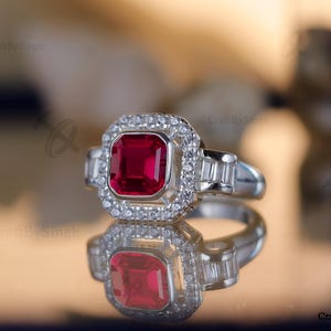 May include: A silver ring with a large, square, ruby-red gemstone at the center, surrounded by a halo of smaller, clear gemstones. The ring's band features additional clear gemstones. The ring is displayed on a reflective surface.