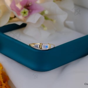 May include: A gold ring with a rectangular, iridescent gemstone center and two baguette-cut side stones. The ring is set in a gold band and is displayed in a teal jewelry box, with flowers in the background. The ring is a piece of jewelry.