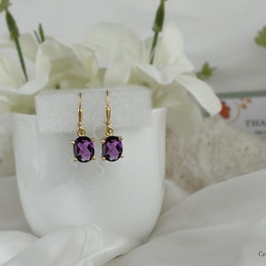 May include: A pair of gold earrings with oval-shaped purple gemstones. The earrings are hanging from a white surface.