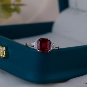 May include: A silver ring featuring a large, rectangular, deep red gemstone. Two smaller, triangular, clear gemstones are either side of the main stone. The ring is displayed in a teal jewellery box with a gold emblem.