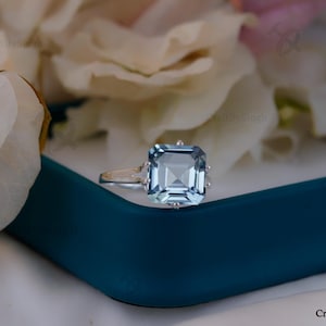 May include: A square-cut, light blue gemstone ring with a silver band. The ring is set on a teal surface, with white and pink flowers in the background. The ring's gemstone is approximately 1.9 cm wide.