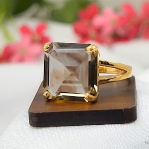 May include: A gold ring with a large, clear, smoky quartz gemstone. The gemstone is cut in a square shape with beveled edges. The ring is set on a wooden display stand.