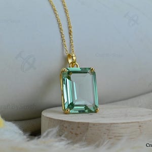 May include: A gold necklace featuring a rectangular, emerald-cut light green gemstone pendant. The pendant is held by a gold-toned metal setting and hangs from a delicate gold chain. The gemstone is translucent and displays a clear, faceted surface.