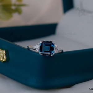 May include: A silver ring featuring a large, square, deep blue gemstone, with two smaller, triangular, clear gemstones either side. The ring is presented in a teal jewellery box with a gold emblem.