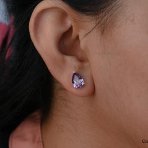 May include: A close-up of a person's ear with a single, teardrop-shaped amethyst stud earring. The earring has a silver setting and a vibrant purple gemstone. The image is well-lit, highlighting the earring's details.