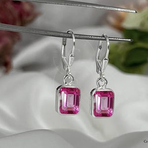 Puede incluir: Pendientes de palanca de plata con piedras preciosas rosas rectangulares. Las piedras están engastadas en un bisel de plata y tienen un tono rosa vibrante. Los pendientes están sujetos por una herramienta plateada sobre un fondo blanco suave.