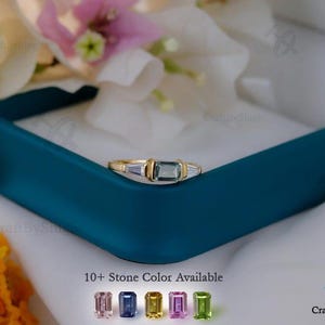 Könnte beinhalten: Ein Goldring mit einem zentralen rechteckigen blauen Edelstein, flankiert von zwei kleineren klaren Steinen im Baguetteschliff. Der Text 10+ Stone Color Available steht über einer Reihe von fünf kleinen rechteckigen Edelsteinen in Rosa, Blau, Gelb, Lila und Grün.