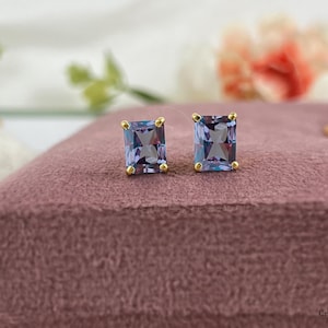 May include: A pair of gold stud earrings with a square-shaped purple gemstone.