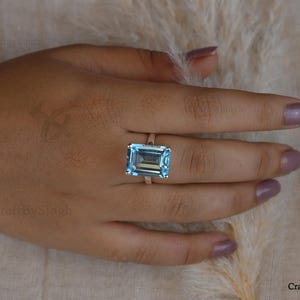 May include: A ring featuring a large, rectangular, emerald-cut light blue gemstone set in a simple metal band. The stone is transparent and faceted, reflecting light across its surface. The band is thin and metallic, encircling the finger.