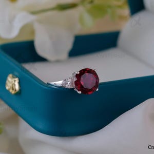 May include: A silver ring with a large, round, deep red gemstone as the centerpiece, flanked by smaller, clear stones. The ring rests in a teal jewelry box with a white satin lining, against a backdrop of white fabric and flowers.