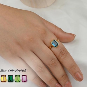 Pode incluir: Um anel de ouro com uma pedra preciosa azul retangular de corte esmeralda em uma cravação de bisel. O texto 10+ Stone Color Available é exibido ao lado de uma fileira de cinco pequenas pedras retangulares em azul claro, amarelo, verde, lima e rosa.