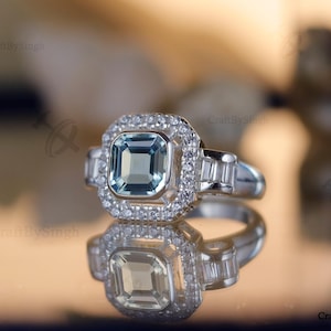 May include: A square-cut, light blue gemstone ring with a halo of small, clear stones. The ring has a silver band and is reflected in a shiny surface. The ring is a piece of jewellery.