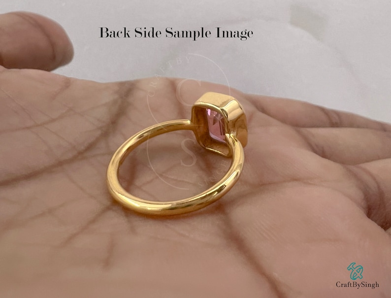 May include: A gold ring with a pink gemstone set in a square shape. The ring is on a hand, but the hand is not visible in the image.