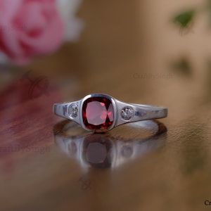May include: A silver ring with a large, square, deep red gemstone in the center, flanked by two smaller, clear stones. The ring is set on a reflective surface, with a blurred floral background.