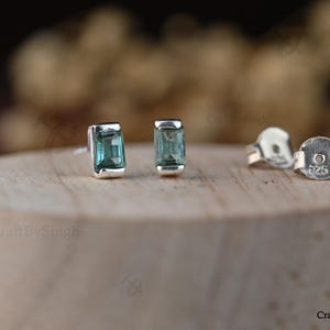 Può includere: Un paio di orecchini a lobo con pietre verdi rettangolari taglio smeraldo incastonate in argento. Gli orecchini includono due chiusure a farfalla in argento, ciascuna chiaramente marchiata con il numero 925, che indica la composizione in argento sterling.